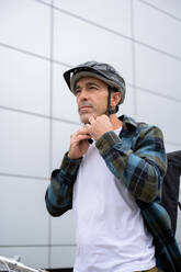 Serious young male in casual clothes and cap touching chin and looking at camera while standing against tiled wall of modern building - ADSF50110