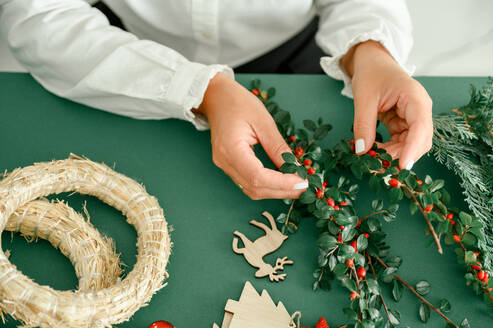 From below of crop anonymous female decorating and holding Christmas wreath with green branches with red berries, green background next to wreath different ornaments, craft stuff. Christmas concept - ADSF50111