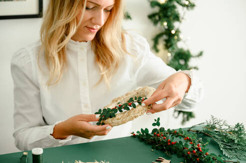 From below of crop anonymous female decorating and holding Christmas wreath with green branches with red berries, green background next to wreath different ornaments, craft stuff. Christmas concept - ADSF50112