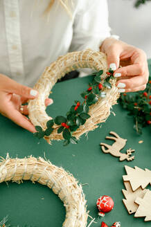 From below of crop anonymous female decorating and holding Christmas wreath with green branches with red berries, green background next to wreath different ornaments, craft stuff. Christmas concept - ADSF50113