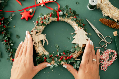 Top view of crop anonymous hand of person holding red and green Christmas wreath placed on table with various Christmas stuff, scissors, ornaments, ribbons - ADSF50114