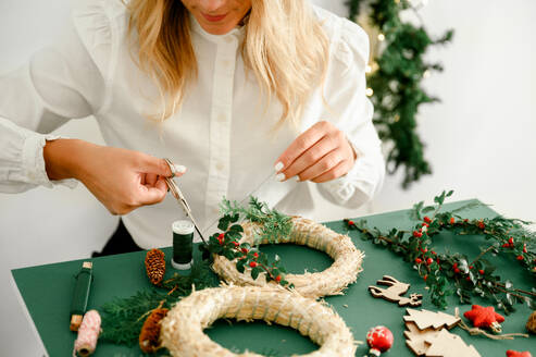 From below of crop anonymous female decorating Christmas wreath with green branches with red berries and baubles on green background next to wreath different ornaments, craft stuff. Christmas content - ADSF50116