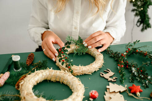 From below of crop anonymous female decorating Christmas wreath with green branches with red berries and baubles on green background next to wreath different ornaments, craft stuff.. Christmas concept - ADSF50117