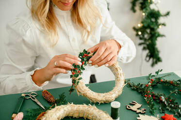 From below of crop anonymous female decorating Christmas wreath with green branches with red berries and baubles on green background next to wreath different ornaments, craft stuff. Christmas content - ADSF50118