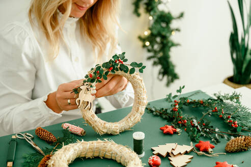 From below of crop anonymous female decorating Christmas wreath with green branches with red berries and baubles on green background next to wreath different ornaments, craft stuff. Christmas concept - ADSF50119