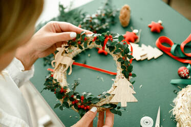 Top view of Christmas wreath, flat lay. Hands holding christmas wreath with branches, berries, pine cones, and ribbon isolated on green background. Christmas crown workshop. Christmas background. concept - ADSF50120