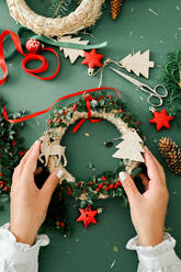 Top view of Christmas wreath, flat lay. Hands holding christmas wreath with branches, berries, pine cones, and ribbon isolated on green background. Christmas crown workshop. Christmas background. - ADSF50121