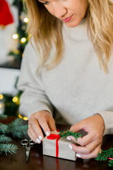 Crop of Young woman in sweater wrapping Christmas presents using paper, scissors, colorful ribbons and decorate present with fir at home. Festive holiday preparations. christmas and new year concept - ADSF50123
