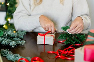 Crop of unrecognizable Woman in sweater wrapping christmas gifts in paper, in background christmas lights next to her green fir and wrapping paper on table - ADSF50124