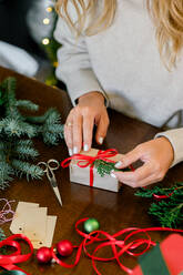 Crop of woman in sweater wrapping Christmas presents using paper, scissors, colorful ribbons and putting fir on present box at home. In Background lights. Christmas concept - ADSF50125