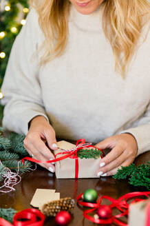 Crop of unrecognizable Woman in sweater holding Christmas present decorated with red ribbon and fir in hand on table, in background Christmas lights next. Christmas and new Year Concept - ADSF50126