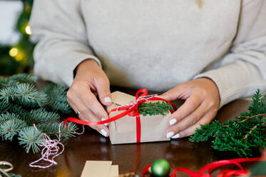 Crop of unrecognizable Woman in sweater holding christmas present decorated with red ribbon and fir in hand, in background christmas lights next to her green fir and wrapping paper on table - ADSF50127