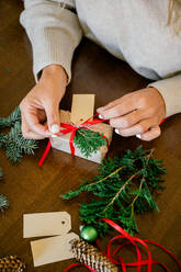 Crop of hand of woman in sweater wrapping Christmas presents using paper, scissors, colorful ribbons and putting fir on present box at home. In Background lights. Christmas concept - ADSF50129