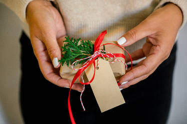 Crop of unrecognizable Woman in sweater holding christmas present decorated with red ribbon and fir in hand, in background christmas lights next to her green fir and wrapping paper on table - ADSF50130