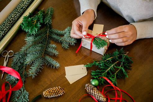 Crop of woman's hand in sweater wrapping Christmas presents using paper, scissors, colorful ribbons and putting fir on present box at home. In Background lights. Christmas concept - ADSF50131