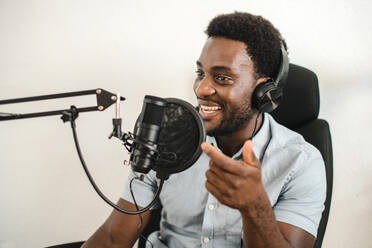 Glad African American male radio host in headphones sitting at table and speaking in microphone while recording podcast in studio on white background - ADSF50138