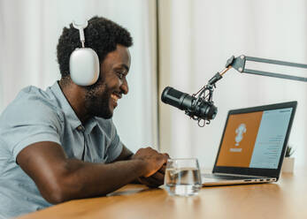 Side view of cheerful male in wireless headphones browsing computer while preparing for recording podcast and sitting at table in studio - ADSF50141