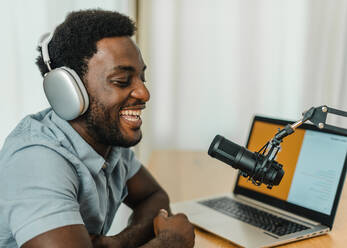 Side view of glad male in headphones speaking in microphone while recording podcast and sitting at table with laptop in studio - ADSF50142