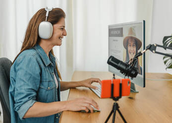 Side view of smiling female influencer in wireless headphones typing on keyboard and looking at screen of computer monitor while recording podcast at table with mic - ADSF50145