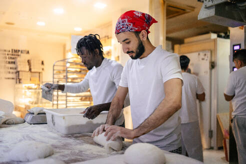 Arabic baker in white clothes and red cap kneading to make bread with a black coworker next to him cutting the dough into pieces in a bakery - ADSF50153