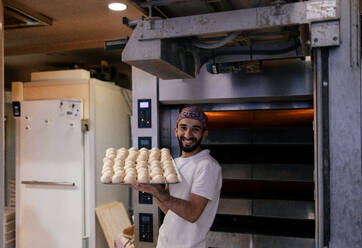 Portrait of a young arabic baker in a white t-shirt and red cap carrying a tray with a multitude of small dough balls smiling in a bakery with the oven in the background - ADSF50154