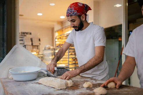 Junger arabischer Bäcker in weißer Kleidung und mit rotem Hut schneidet Teig mit einem Skapulier in einer Bäckerei - ADSF50155