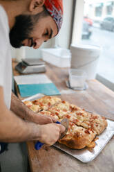 Young arabic pizza maker in white t-shirt and red hat cutting pizza in slices with a cutter in a pizzeria - ADSF50156
