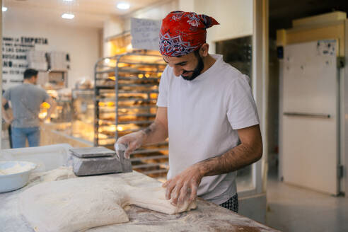 Junger arabischer Bäcker in weißer Kleidung und mit rotem Hut schneidet Teig mit einem Schulterblatt und wiegt ihn auf einer Waage in einer Bäckerei - ADSF50157