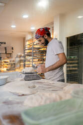 Junger arabischer Bäcker in weißer Kleidung und mit rotem Hut schneidet Teig mit einem Schulterblatt und wiegt ihn auf einer Waage in einer Bäckerei - ADSF50158