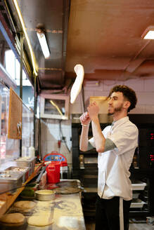 Portrait of young arabic pizza man in white apron throwing the dough in the air in a pizzeria - ADSF50160