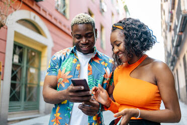 Young black couple in colorful and casual clothes walking around the city while using their smartphone in the street. The man shows something to the woman on the smartphone while smiling - ADSF50161