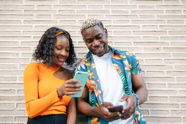 Young black couple in colorful and casual clothes leaning against the wall while both use their smartphone smiling. The woman is showing something in her smartphone to the man - ADSF50163