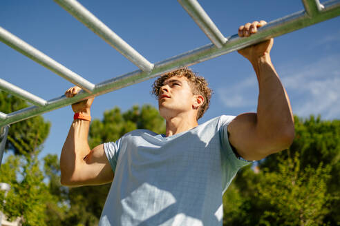 Handsome young man with blond hair dressed in sportswear doing pull ups in an outdoor calisthenics park on a sunny day - ADSF50167