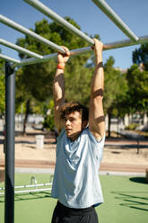 Handsome young man with blond hair dressed in sportswear doing pull ups in an outdoor calisthenics park on a sunny day - ADSF50169