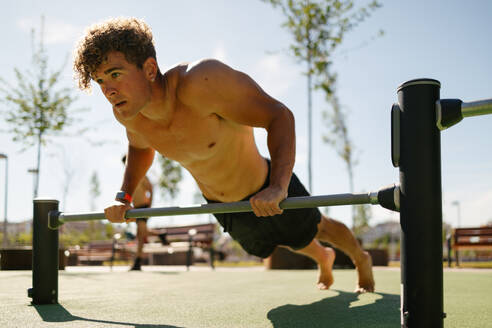 Handsome young man with blond hair shirtless doing barbell push-ups in an outdoor calisthenics park on a sunny day - ADSF50173