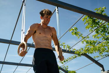 Handsome young man with blond hair shirtless exercising with rings outdoors on sunny day - ADSF50174