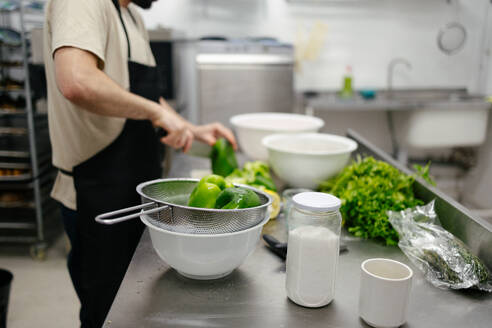 Close-up of green peppers while in the background there is an out-of-focus and unrecognizable chef cutting peppers in a professional kitchen - ADSF50176