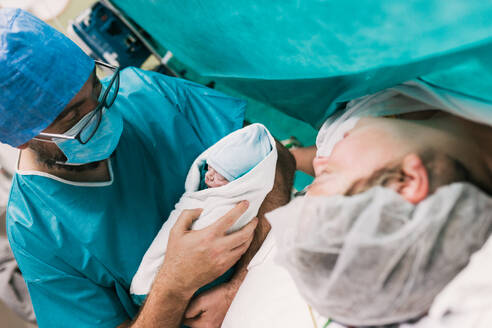 High angle of male obstetrician in medical uniform and glasses demonstrating infant in blanket to mom after C section surgery in hospital - ADSF50179