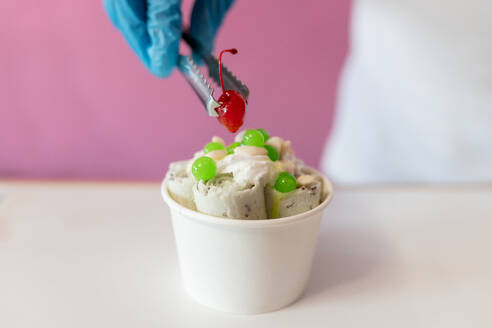 A worker is decorating a rolled ice cream tub with a cherry on top using tongs. Concept of new small business - ADSF50182