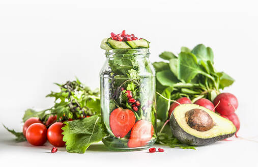 Green salad in jar with ingredients: green lettuce leaves, tomato, strawberry, avocado, cucumber and pomegranate seeds at white background. Healthy to go food with vegetables and fruits. Front view. - ADSF50188