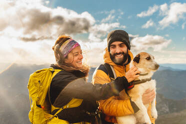 Couple of hikers in the rain making a mountain route with their dog. Mountaineer holding his dog in his arms with a mountain range landscape in the background. woman petting dog - ADSF50213