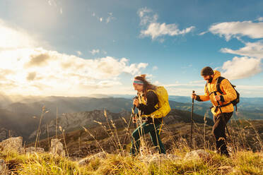 Zwei Bergsteiger, ein Mann und eine Frau, wandern bei Sonnenuntergang auf einen Berg. Die Wanderer sind mit Rucksäcken, Trekkingstöcken und warmer Kleidung ausgestattet und wandern den Berg hinauf. - ADSF50215