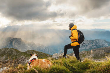 Unbekannte Person mit Rucksack, die auf einem Felsen sitzt und die Landschaft betrachtet, nachdem sie mit ihrem Hund einen Berg bestiegen hat. Wanderer, der an einem bewölkten Tag den Gipfel erreicht. - ADSF50221