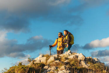 Man and woman with backpacks and trekking poles contemplating the sunset from the top of a mountain. two hikers summiting a mountain peak. - ADSF50224