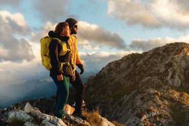 Couple of hikers equipped with backpacks and abirgo clothing, standing on a mountain peak watching the sunset with their dog. outdoor sports and healthy lifestyle - ADSF50225