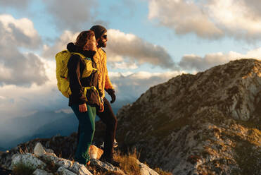 Ein Paar Wanderer, ausgerüstet mit Rucksäcken und Abirgo-Kleidung, stehen auf einem Berggipfel und beobachten den Sonnenuntergang mit ihrem Hund. Outdoor-Sport und gesunder Lebensstil - ADSF50225