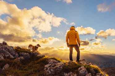 Unbekannte Person mit Rucksack, die auf einem Felsen sitzt und die Landschaft betrachtet, nachdem sie mit ihrem Hund einen Berg bestiegen hat. Wanderer, der an einem bewölkten Tag den Gipfel erreicht. - ADSF50228