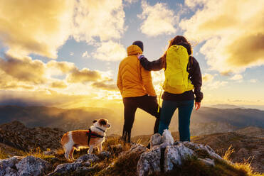 Ein Wanderpärchen mit ihrem Hund, das den Sonnenuntergang vom Gipfel eines Berges aus beobachtet. Sport und Aktivitäten in den Bergen und im Freien. - ADSF50229