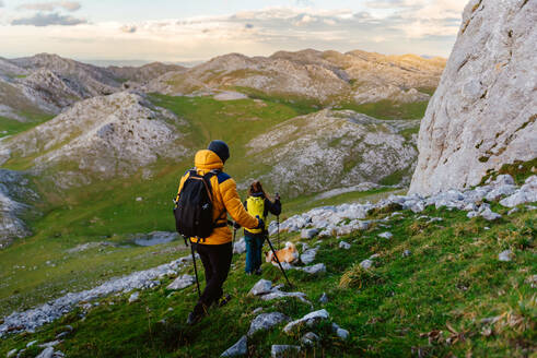 Ein Paar Wanderer mit ihrem Hund, die ein grünes Tal und die Berge betrachten. Bergsteiger, die mit Trekkingstöcken und Rucksäcken einen Berg hinabsteigen. Outdoor-Sport. - ADSF50232