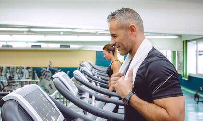 Portrait of smiling man with towel in his neck training over a treadmill on fitness center - ADSF50242